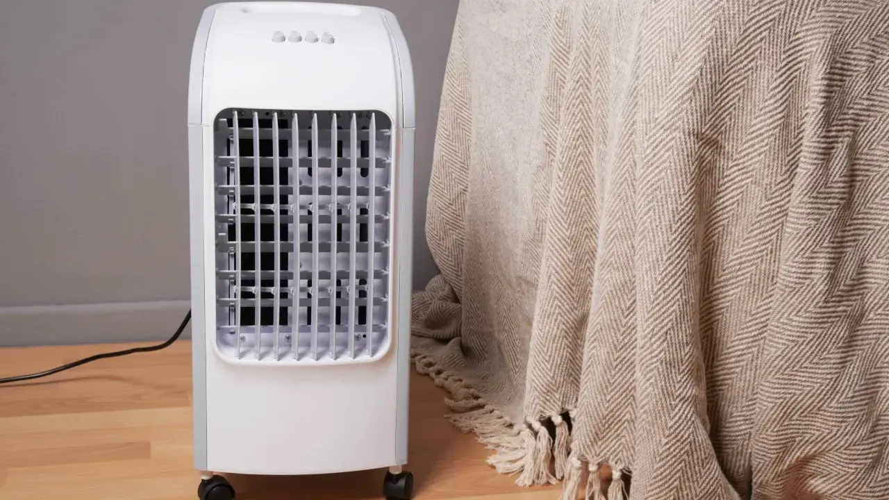 Un rafraîchisseur d'air blanc sur roulettes, avec une grille frontale, à côté d'une couverture beige à chevrons.