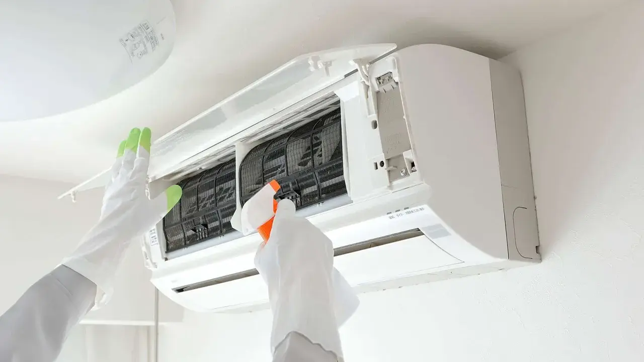 Gants blancs et verts pour l'entretien climatisation. Une main vaporise un produit sur les filtres d'un climatiseur mural ouvert.