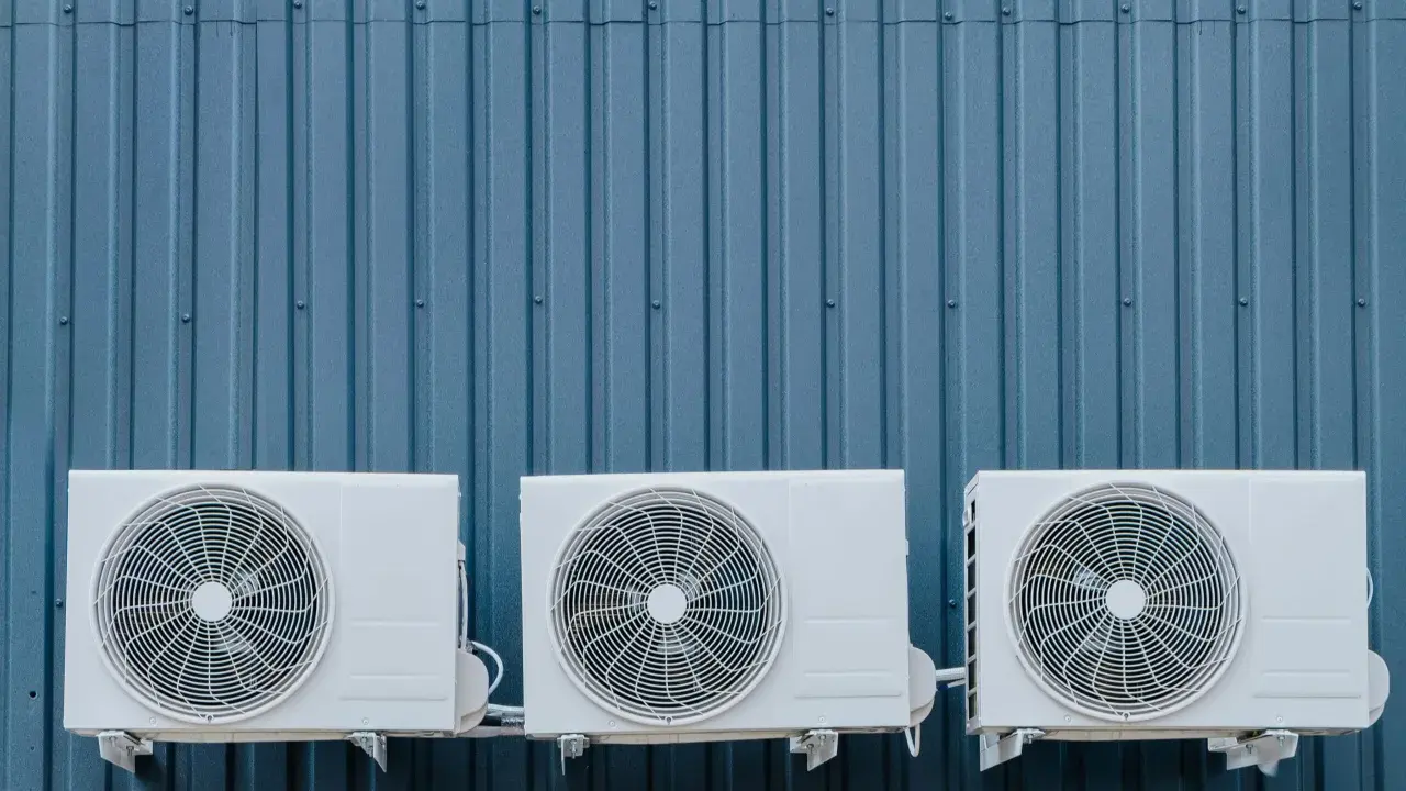 Trois unités extérieures de climatisation blanches sont fixées sur un mur bleu ondulé. La hauteur de ces unités est standard pour une installation extérieure.