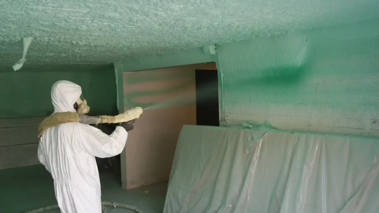 Un ouvrier en combinaison blanche projette du polyuréthane sur un plafond, créant une isolation texturée.