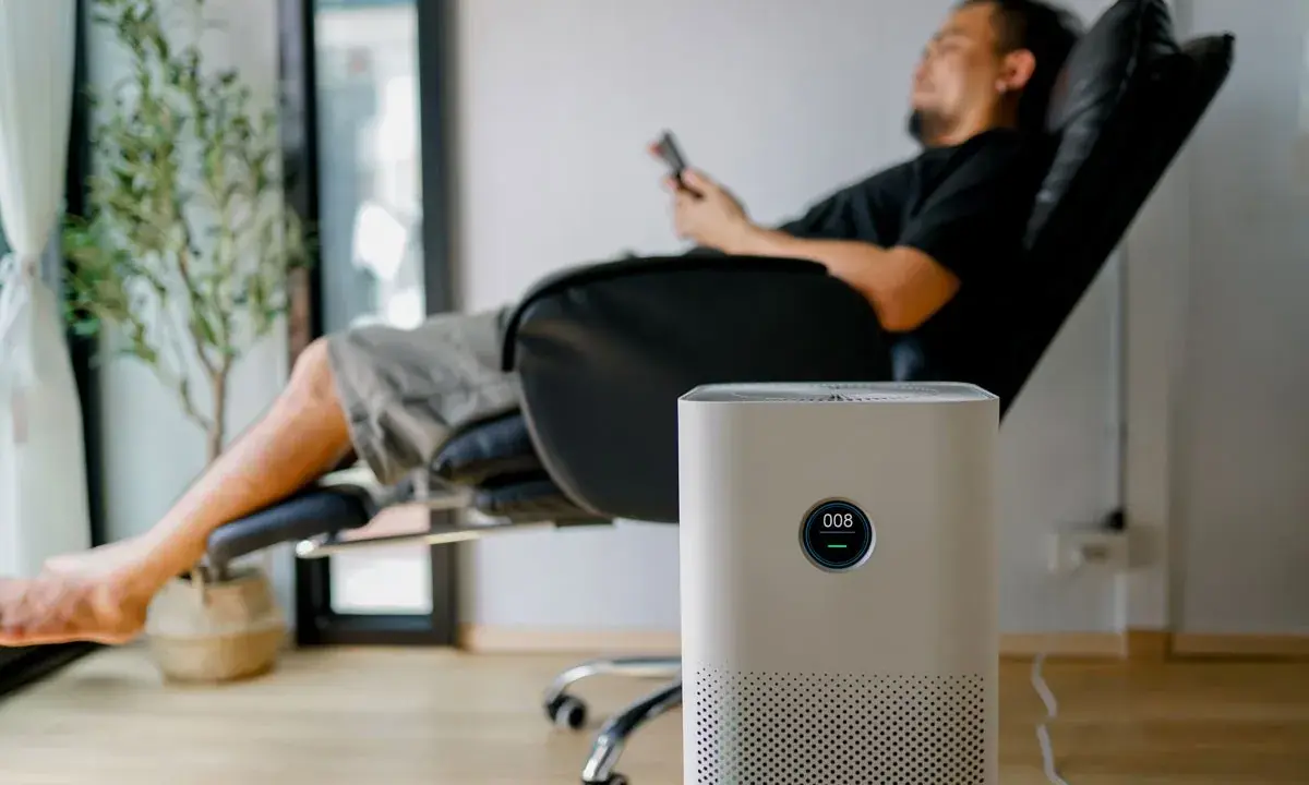 Un homme se repose dans un fauteuil, une plante verte à côté. Un purificateur d'air est au premier plan. Peut-on coucher une clim mobile ? Non, cet appareil est conçu pour rester debout.