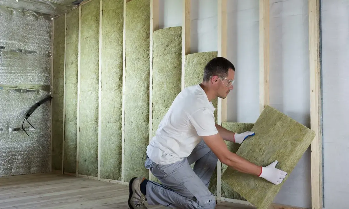 Un ouvrier installe des panneaux d'isolant minéral pour le **cout isolation interieur**. Le mur est en cours de construction, avec des montants en bois et une isolation déjà en place.