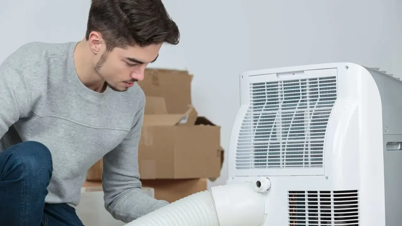 Un jeune homme installe un climatiseur mobile 40m2. Des cartons sont empilés derrière lui.