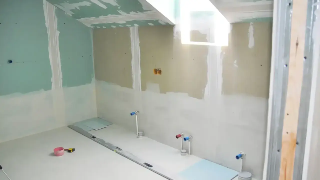 Chantier de salle de bain en cours : murs en plaques de plâtre, arrivées d'eau apparentes, niveau à bulle et outils au sol. L'isolation salle de bain est en phase de préparation.