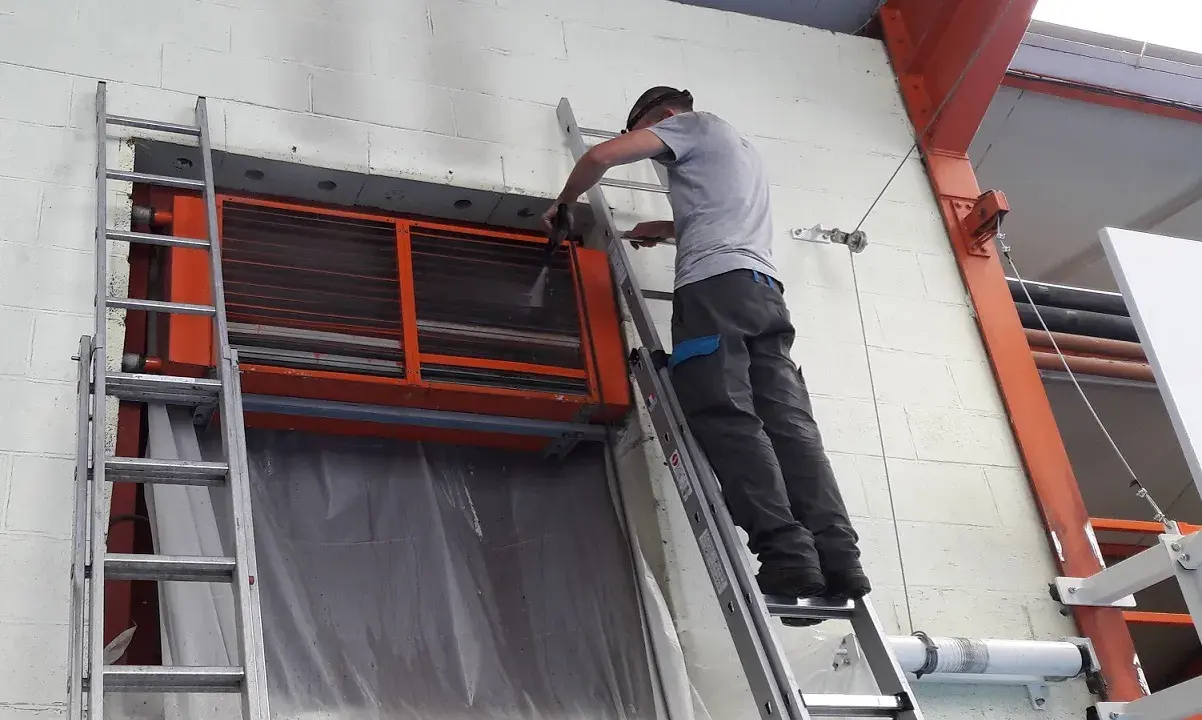 Un technicien sur une échelle nettoie un aérotherme de chauffage industriel.