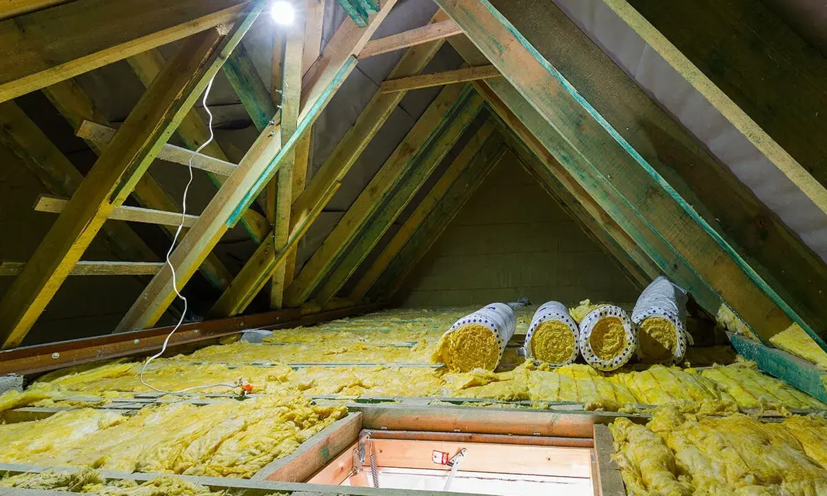 Travaux en cours pour isoler les combles. Rouleaux d'isolant jaune posés sur la structure en bois, sous un éclairage vif.