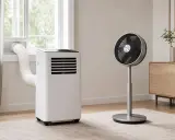 Un climatiseur sans évacuation, avec son tuyau dirigé vers la fenêtre, et un ventilateur sur pied dans une pièce lumineuse.