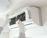 Gants blancs et verts pour l'entretien climatisation. Une main vaporise un produit sur les filtres d'un climatiseur mural ouvert.
