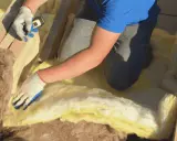 Pose d'isolant jaune entre les solives pour l'isolation plancher bas. Gants blancs et bleus, mètre en main.