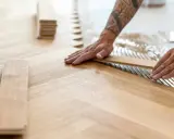 Pose de lames de parquet en bois, créant un motif en chevrons. L'isolation du sol est assurée par la colle appliquée.