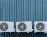 Trois unités extérieures de climatisation blanches sont fixées sur un mur bleu ondulé. La hauteur de ces unités est standard pour une installation extérieure.