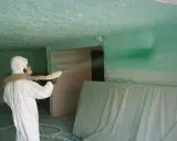 Un ouvrier en combinaison blanche projette du polyuréthane sur un plafond, créant une isolation texturée.