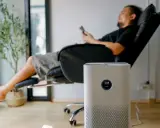 Un homme se repose dans un fauteuil, une plante verte à côté. Un purificateur d'air est au premier plan. Peut-on coucher une clim mobile ? Non, cet appareil est conçu pour rester debout.