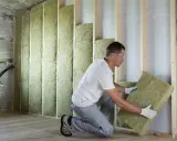 Un ouvrier installe des panneaux d'isolant minéral pour le **cout isolation interieur**. Le mur est en cours de construction, avec des montants en bois et une isolation déjà en place.