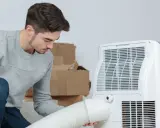 Un jeune homme installe un climatiseur mobile 40m2. Des cartons sont empilés derrière lui.
