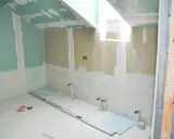 Chantier de salle de bain en cours : murs en plaques de plâtre, arrivées d'eau apparentes, niveau à bulle et outils au sol. L'isolation salle de bain est en phase de préparation.