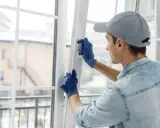 Un ouvrier ajuste une fenêtre pour isoler du froid. Il porte des gants bleus et une casquette grise.