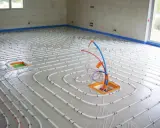 Installation de tubes pour un chauffage électrique au sol. Le sol est recouvert d'une bâche et les tubes blancs sont disposés en spirale.