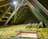 Travaux en cours pour isoler les combles. Rouleaux d'isolant jaune posés sur la structure en bois, sous un éclairage vif.