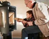 Un grand-père aide son petit-fils à charger du bois dans un poêle. Une alternative au chauffage au fioul, mais attention aux dangers du feu.