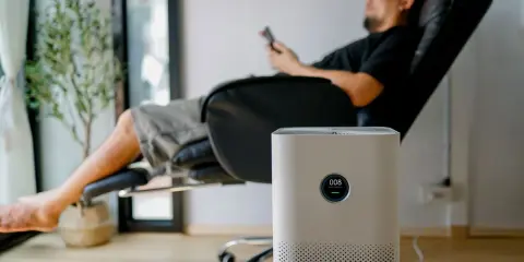 Un homme se repose dans un fauteuil, une plante verte à côté. Un purificateur d'air est au premier plan. Peut-on coucher une clim mobile ? Non, cet appareil est conçu pour rester debout.