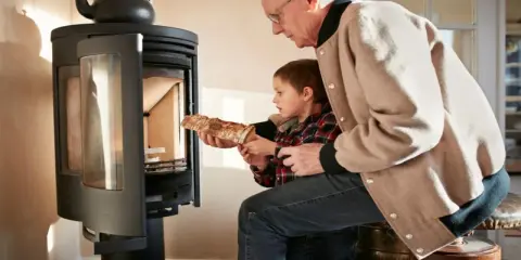 Un grand-père aide son petit-fils à charger du bois dans un poêle. Une alternative au chauffage au fioul, mais attention aux dangers du feu.