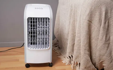 Un rafraîchisseur d'air blanc sur roulettes, avec une grille frontale, à côté d'une couverture beige à chevrons.