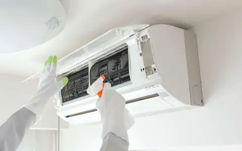 Gants blancs et verts pour l'entretien climatisation. Une main vaporise un produit sur les filtres d'un climatiseur mural ouvert.