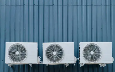 Trois unités extérieures de climatisation blanches sont fixées sur un mur bleu ondulé. La hauteur de ces unités est standard pour une installation extérieure.
