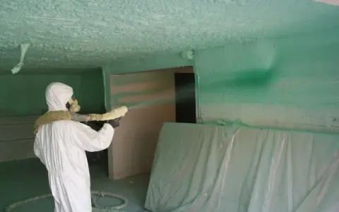 Un ouvrier en combinaison blanche projette du polyuréthane sur un plafond, créant une isolation texturée.