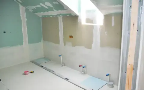 Chantier de salle de bain en cours : murs en plaques de plâtre, arrivées d'eau apparentes, niveau à bulle et outils au sol. L'isolation salle de bain est en phase de préparation.