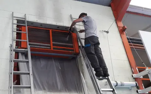 Un technicien sur une échelle nettoie un aérotherme de chauffage industriel.
