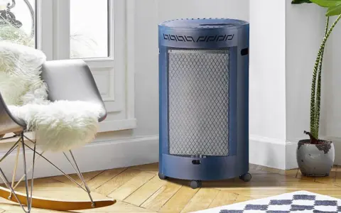 Un chauffage gaz infrarouge ou infrableu bleu, cylindrique et sur roulettes, diffuse sa chaleur dans un salon cosy, près d'un fauteuil confortable.