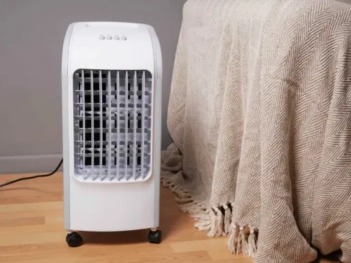 Un rafraîchisseur d'air blanc sur roulettes, avec une grille frontale, à côté d'une couverture beige à chevrons.