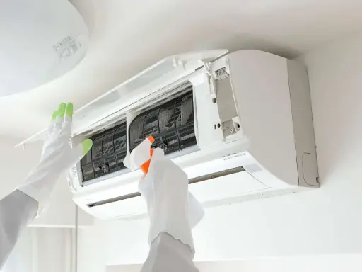 Gants blancs et verts pour l'entretien climatisation. Une main vaporise un produit sur les filtres d'un climatiseur mural ouvert.