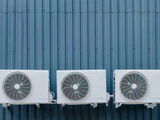 Trois unités extérieures de climatisation blanches sont fixées sur un mur bleu ondulé. La hauteur de ces unités est standard pour une installation extérieure.
