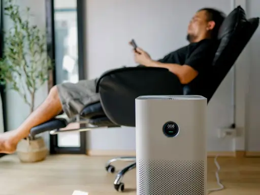 Un homme se repose dans un fauteuil, une plante verte à côté. Un purificateur d'air est au premier plan. Peut-on coucher une clim mobile ? Non, cet appareil est conçu pour rester debout.
