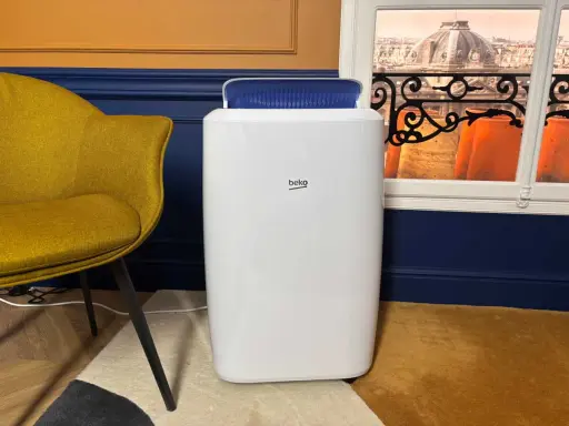 Climatiseur Beko, blanc, avec une sortie d'air bleue. Un fauteuil moutarde et une vue sur des toits parisiens. Avis positifs attendus.