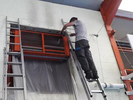 Un technicien sur une échelle nettoie un aérotherme de chauffage industriel.