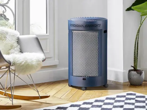 Un chauffage gaz infrarouge ou infrableu bleu, cylindrique et sur roulettes, diffuse sa chaleur dans un salon cosy, près d'un fauteuil confortable.