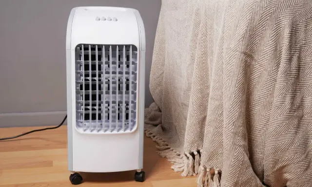Un rafraîchisseur d'air blanc sur roulettes, avec une grille frontale, à côté d'une couverture beige à chevrons.