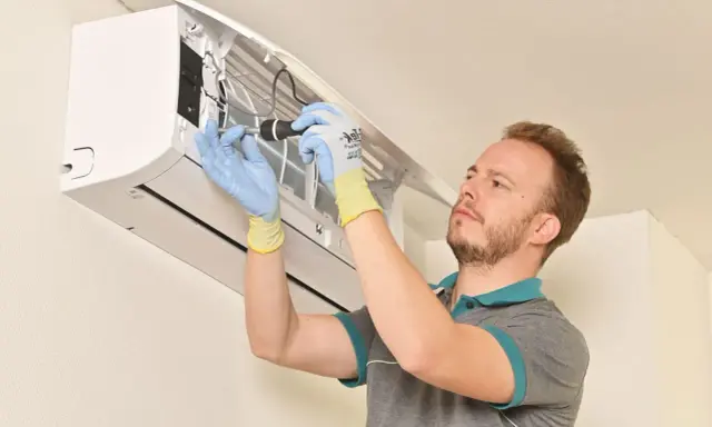 Technicien en gants bleus et jaunes effectuant la mise en service d'une clim split, outil à la main.