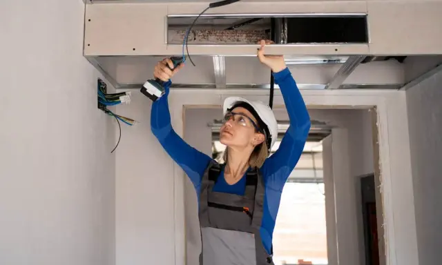 Installation d'un système de climatisation gainable plafond. Une ouvrière visse une plaque de plâtre.