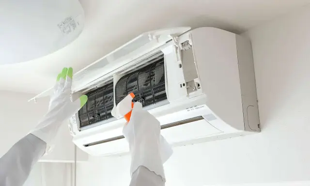 Gants blancs et verts pour l'entretien climatisation. Une main vaporise un produit sur les filtres d'un climatiseur mural ouvert.