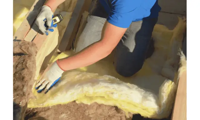 Pose d'isolant jaune entre les solives pour l'isolation plancher bas. Gants blancs et bleus, mètre en main.