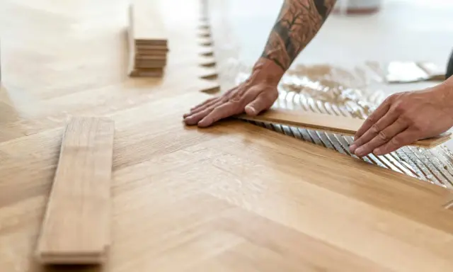 Pose de lames de parquet en bois, créant un motif en chevrons. L'isolation du sol est assurée par la colle appliquée.