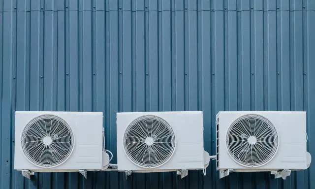 Trois unités extérieures de climatisation blanches sont fixées sur un mur bleu ondulé. La hauteur de ces unités est standard pour une installation extérieure.