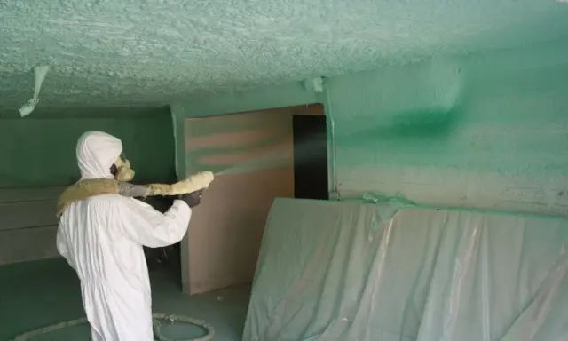 Un ouvrier en combinaison blanche projette du polyuréthane sur un plafond, créant une isolation texturée.