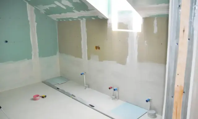 Chantier de salle de bain en cours : murs en plaques de plâtre, arrivées d'eau apparentes, niveau à bulle et outils au sol. L'isolation salle de bain est en phase de préparation.