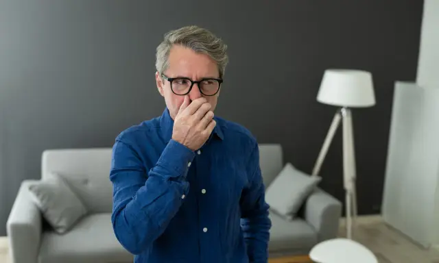 Homme d'âge mûr se pinçant le nez, visiblement incommodé par une mauvaise odeur de clim neuve dans son salon moderne.